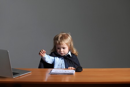 Little boy child smiling in black academic mantle sitting at school wooden desk near notebook computer mouse and diaryの写真素材
