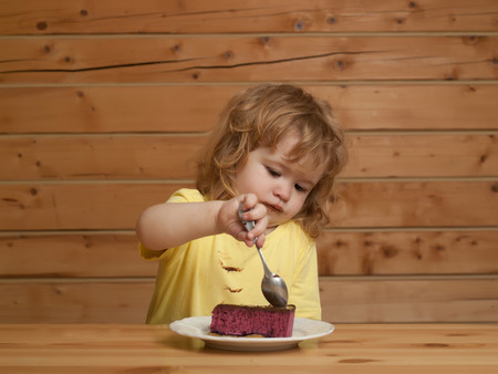 Cute little boy child with long blond hair eats fruit cake pie with chocolate at wooden tableの写真素材