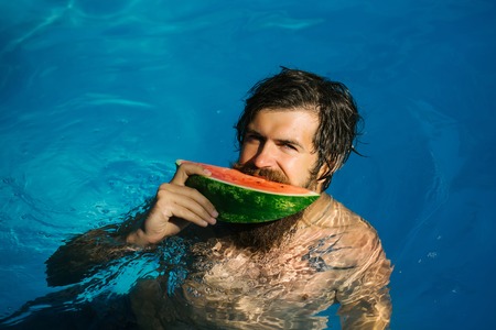 young bearded man with wet hair and beard swimming in pool with blue water holds green red watermelon sunny summer day outdoorの写真素材