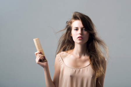 young woman with slim body pretty face and brunette long hair in dress holding hairbrush standing in studio on grey backgroundの写真素材