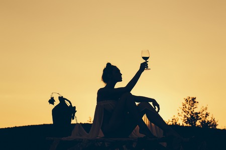 Silhouette of slim girl young woman female model sommelier with wine glass and wicker bottle on nature over beautiful sunsetの写真素材