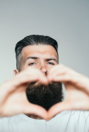 handsome young man with long beard and moustache on face holding hands in heart shape on grey background in studioの写真素材