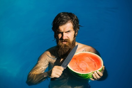 young sexy bearded man with wet hair and beard swimming in pool with blue water holds knife and green red watermelon sunny summer day outdoorの写真素材