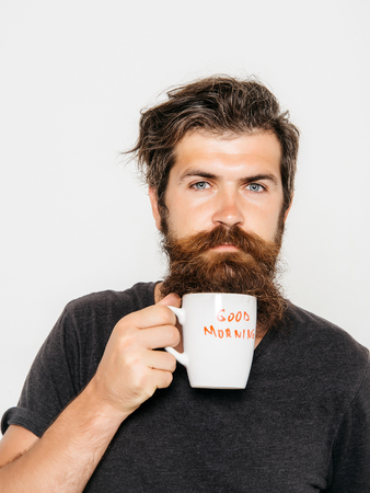 handsome bearded man with stylish hair beard and mustache on serious face in shirt holding white cup or mug with good morning text drinking tea or coffee in studio on grey backgroundの写真素材