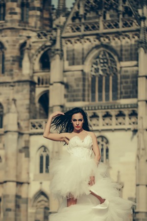 young sexy girl with brunette hair and red lips on pretty face in white wedding bride dress posing near castle or palace building sunny outdoorの写真素材
