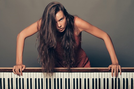 young girl or woman with pretty face and long brunette hair has body posing near old retro wooden piano with keyboard on grey backgroundの写真素材