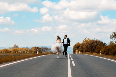 Beautiful bride and handsome groom elegant fashion married couple in wedding dress and suit run along road and hold hands on blue skyの写真素材