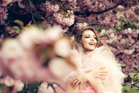 Beautiful young woman in pink glamour dress with long curly hair and smiling face in spring flowers bloomの写真素材