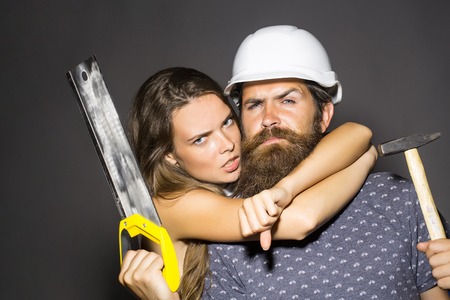 Young woman with pretty face hugging man repairman with bearded serious face in white building helmet holding hard hammer and headsaw instrument studio on grey backgroundの写真素材