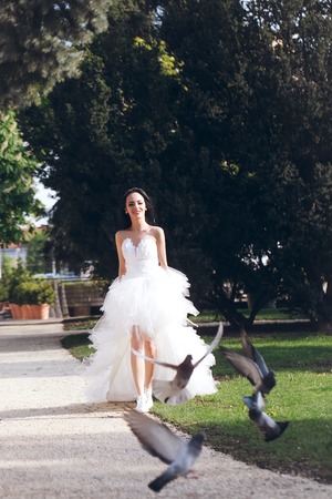 young girl or woman with happy pretty face in white wedding bride dress running in green park near dove or pigeon birds sunny outdoorの写真素材