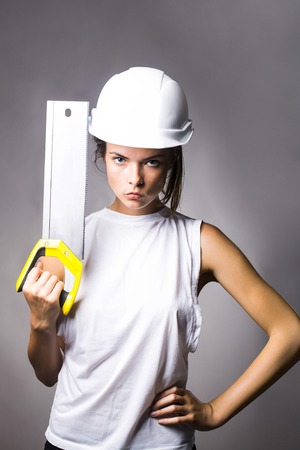 Young girl workwoman repairer with pretty serious strict face in white building helmet holding professional saw instrument in hand studio on gray backgroundの写真素材