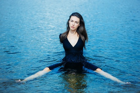 young girl in navy dress with long dark hair sits in water with spread legsの写真素材