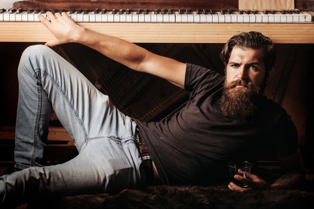 handsome bearded man with stylish hair mustache and beard on serious face drinking brandy or whiskey from glass near old wooden open piano with keyboard as musician with music book, copy spaceの写真素材