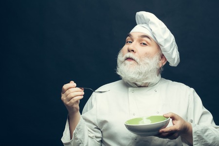 Bearded man cook in hat tasting food with spoon in studio on grey background, copy spaceの写真素材
