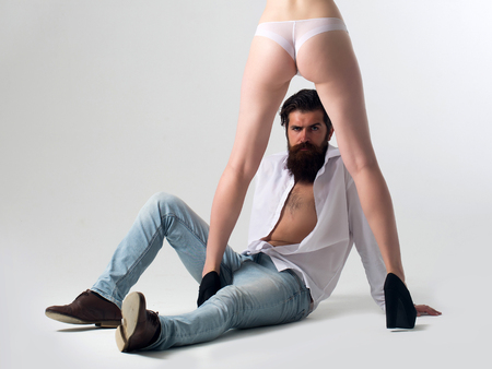handsome bearded man with long beard on serious face sitting near sexy female legs and buttocks in lingerie and shoes of young pretty girl in studio isolated on white background, copy spaceの写真素材