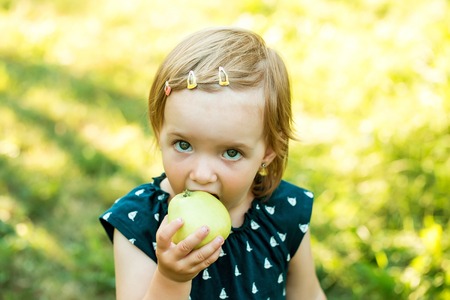 Cute baby girl in blue dress eats apple on sunny summer day on natural backgroundの写真素材
