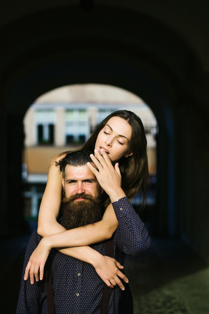 young stylish couple of handsome bearded man with long beard in shirt and pretty sexy woman or girl with brunette hair smoking cigarette embracing in street outdoor with archの写真素材