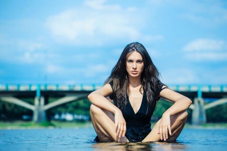 young sexy woman or girl with long hair and pretty face in black summer dress sitting in river or sea water sunny day outdoor on natural backgroundの写真素材