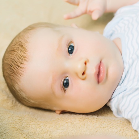 Cute baby boy blond infant with blue eyes in romper lies on his back on beige bed coverの写真素材