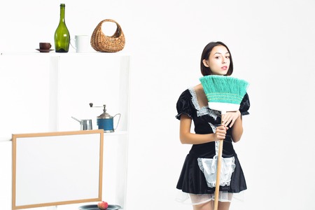 Young girl brunette with pretty face in sexy black maid costume posing with sweep green broom on background of shelves with cookware isolatedの写真素材