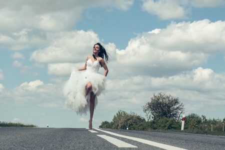 young wedding happy sexy girl or woman with brunette hair and pretty face in white bride dress running on road way on cloudy blue sky backgroundの写真素材