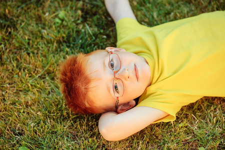 Happy baby boy with red hair in yellow shirt and eyeglasses on cute face laying on green grass summer day outdoor on natural backgroundの写真素材