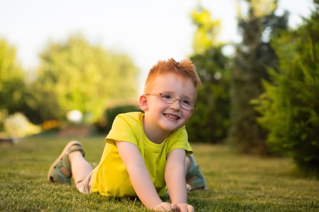 Happy baby boy with red hair in yellow shirt and eyeglasses on happy smiling face laying on green grass summer day outdoor on natural backgroundの写真素材