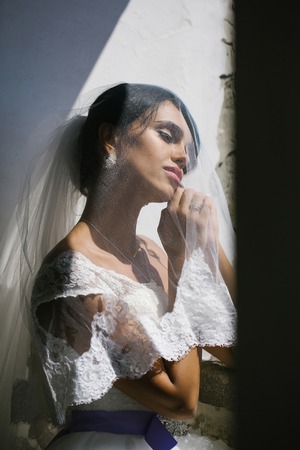 Beautiful bride woman with eyes closed under veil in white wedding dress dreams on sunny day over old wallの写真素材