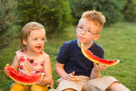 Happy children boy and girl with happy smiling face eating red watermelon summer day outdoor on green grass backgroundの写真素材