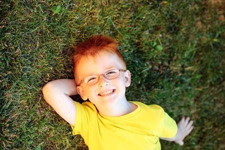 Happy baby boy with red hair in yellow shirt and eyeglasses on happy smiling face laying on green grass summer day outdoor on natural backgroundの写真素材
