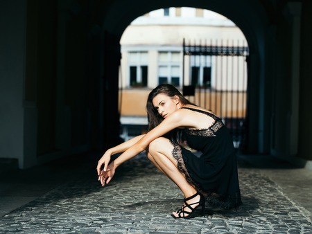 young stylish pretty sexy woman or girl with brunette hair in black dress sitting on paving stones road in street outdoor with archの写真素材