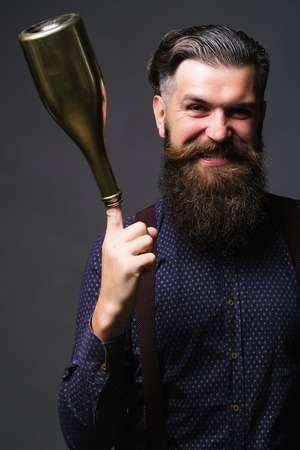 Trendy man with long beard hipster in blue shirt with suspenders holds golden champagne bottle on...