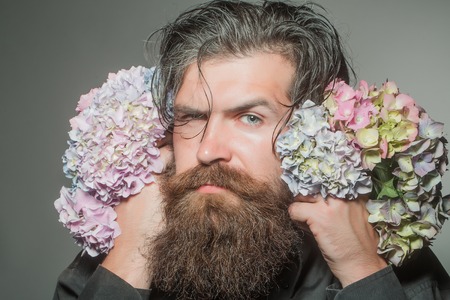 handsome sexy bearded young man hipster with floral beard and mustache on serious face in studio holding hydrangea flower bouquet on grey background, closeupの写真素材