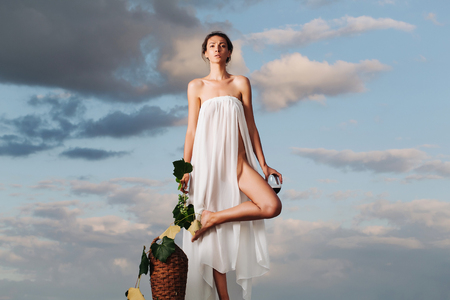 Pretty young girl or woman in white sexy dress denuded with red wine in glass wicker bottle vine and grapes on nature over blue skyの写真素材