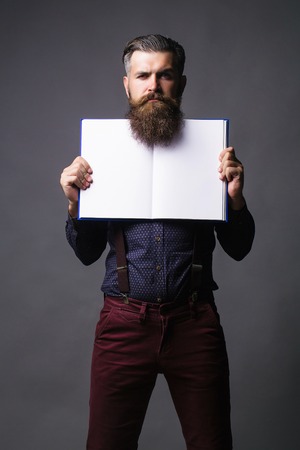 Handsome man with long beard hipster in shirt with suspenders holds open journal for copy space on grey backgroundの写真素材