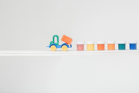 Plastic toy truck with small jars of colorful gouache standing in line on white background
