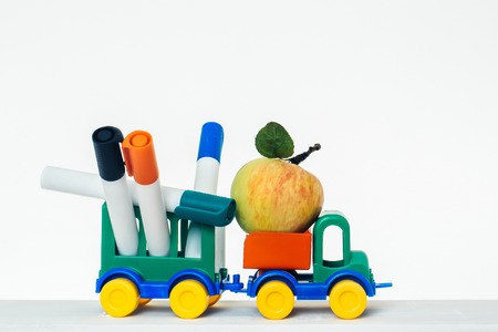 Plastic colorful toy car with one ripe apple and trailer full of markers on white studio background