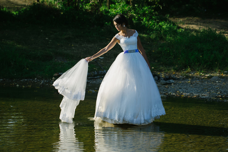 Beautiful bride pretty woman in long elegant white lace wedding dress with veil in hand stands on lake on natural backgroundの写真素材