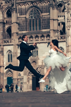 young wedding happy couple of sexy girl with brunette hair and pretty face in white bride dress and handsome man in black groom suit jumping near castle or palace building sunny outdoorの写真素材