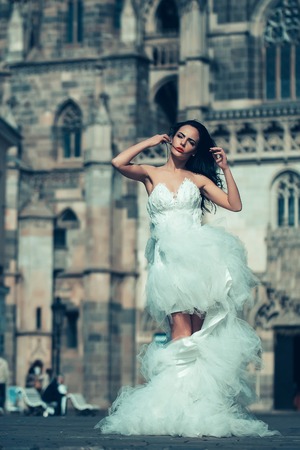 young sexy girl with brunette hair and red lips on pretty face in white wedding bride dress posing near castle or palace building sunny outdoorの写真素材