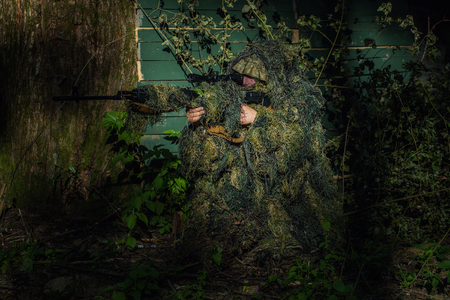 Sniper soldier in ghillie suit camo and mask sitting with rifle aiming target near tree and green branches backgroundの写真素材