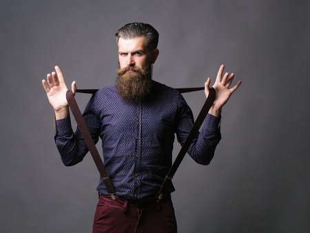Trendy man handsome hipster with long beard and moustache in blue shirt pulls suspenders on grey backgroundの写真素材