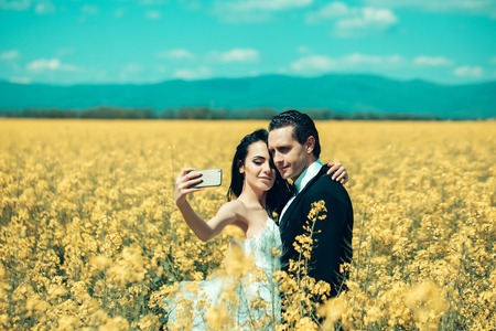 young wedding couple of sexy girl with pretty face in white bride dress holds mobile phone and handsome man in black groom suit in field with yellow flowers on natural background with blue skyの写真素材