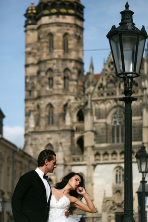 young wedding couple of sexy girl with brunette hair and pretty face in white bride dress and handsome man in black groom suit standing near street lamp on castle or palace building sunny outdoorの写真素材