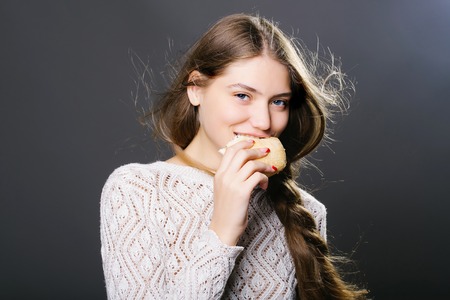 Sexy fashionable girl or woman with pretty smiling face slim model with stylish long brunette hair in white sweater eating bread roll in studio on grey backgroundの写真素材
