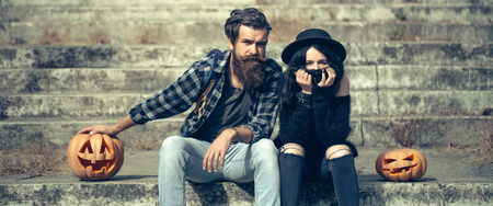 young halloween couple of bearded man with beard and mustache and girl in black witch hat in jeans sit on stony stairs with traditional autumn holiday symbol of orange spooky pumpkin sunny outdoorの写真素材