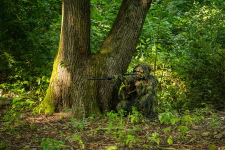 Sniper soldier man in military ghillie suit camouflage with rifle in hands aiming target sitting in ambuscade near tree in forestの写真素材