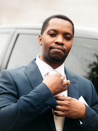 Handsome man african American groom in elegant suit coat for wedding ceremony fixes white tie outdoorsの写真素材