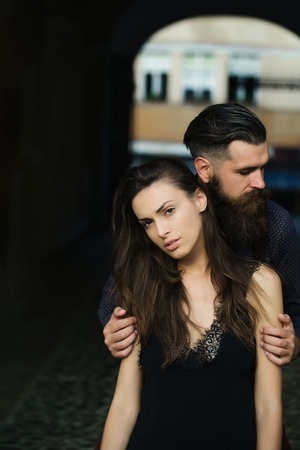 young stylish couple of handsome bearded man with long beard and pretty sexy woman or girl with brunette hair in dress embracing in street outdoor with archの写真素材