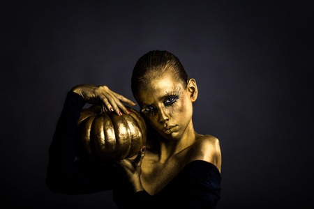 halloween golden woman or girl holding painted gold pumpkin has pretty face with makeup and body art metallized color with bare shoulders on grey backgroundの写真素材